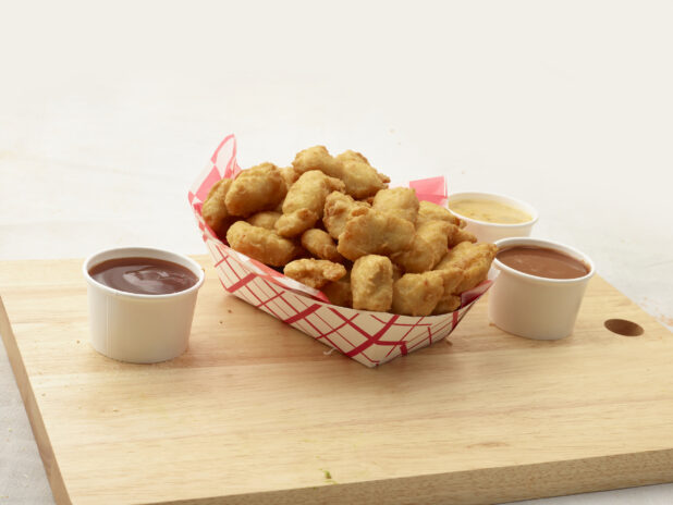 Battered chicken nuggets in a red and white cardboard takeout container, dipping sauces alongside, on a wood board, white background