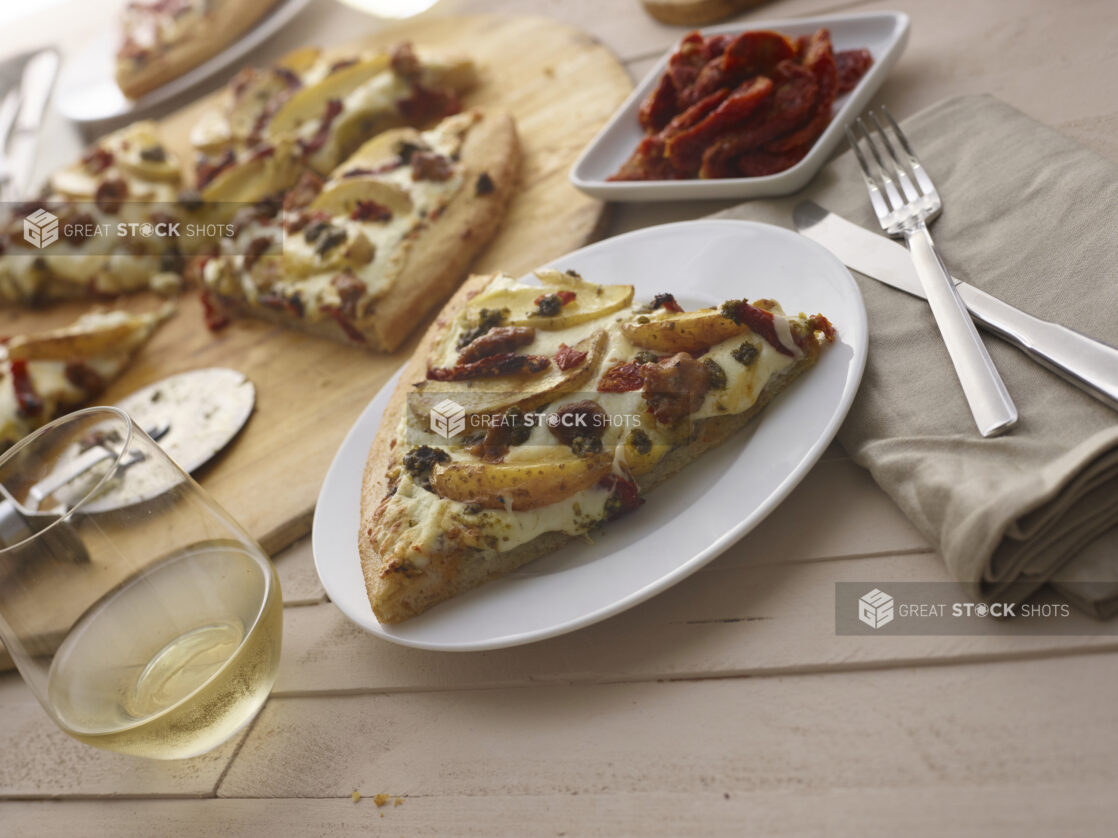 Slice of pizza with potato wedges, sundried tomatoes and sausage on a white side plate, utensils and glass of white wine alongside, pizza and sides in background