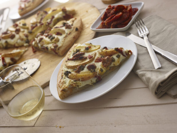 Slice of pizza with potato wedges, sundried tomatoes and sausage on a white side plate, utensils and glass of white wine alongside, pizza and sides in background