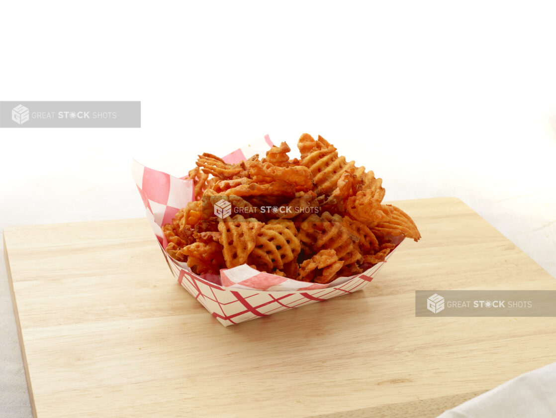 Spicy waffle french fries in a red and white lined cardboard takeout container on a wood board, white background