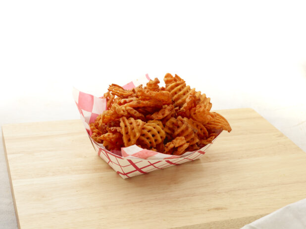 Spicy waffle french fries in a red and white lined cardboard takeout container on a wood board, white background