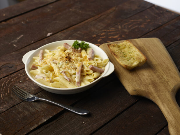 Penne Alfredo with pork belly in a white bowl with handles, side of garlic bread on wood board in background, dark wood background