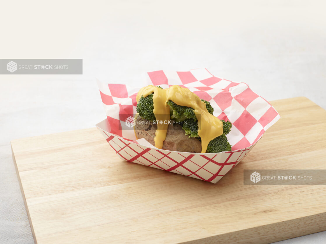 Baked potato stuffed with broccoli and covered in cheddar cheese sauce in a red and white lined cardboard takeout container on a wood board, white background