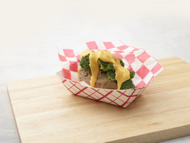 Baked potato stuffed with broccoli and covered in cheddar cheese sauce in a red and white lined cardboard takeout container on a wood board, white background