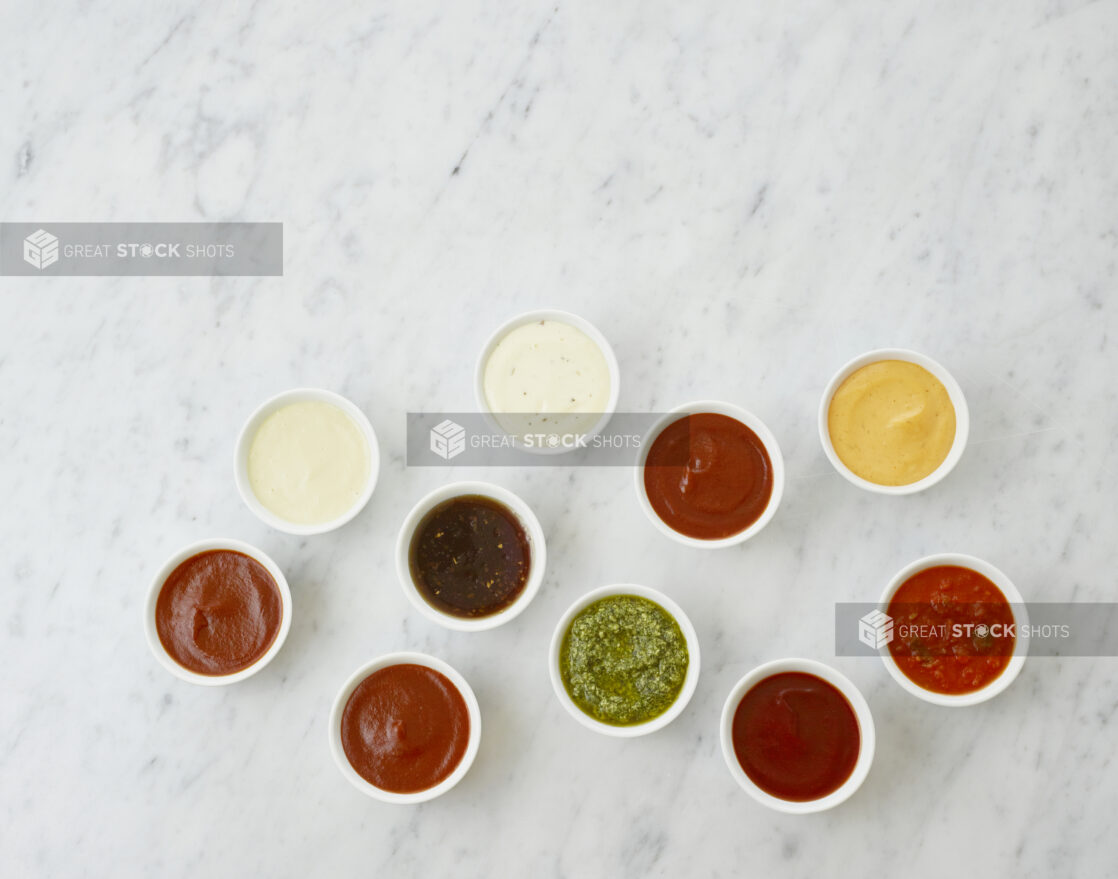 Ten round white ramekins of colourful dipping sauces and dressings, and condiments, overhead view, white marble background