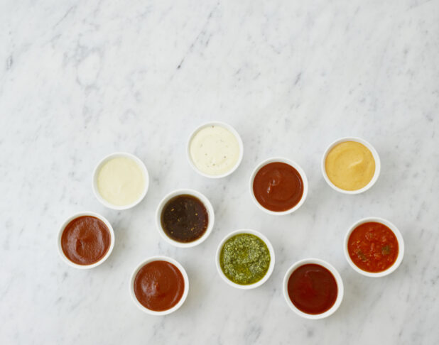 Ten round white ramekins of colourful dipping sauces and dressings, and condiments, overhead view, white marble background