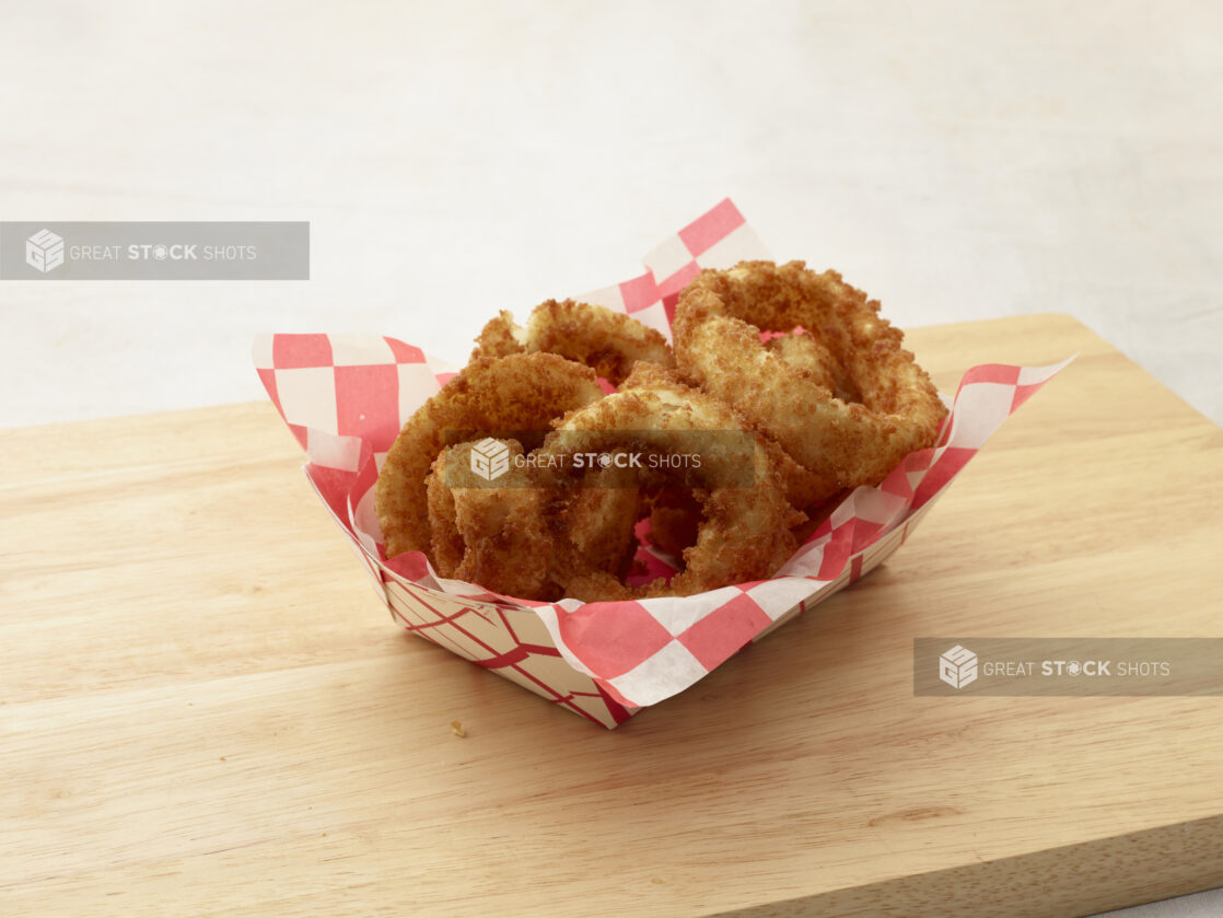 Onion rings in a cardboard takeout container with red and white checkered liner on a wood board, white background