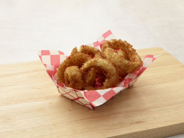 Onion rings in a cardboard takeout container with red and white checkered liner on a wood board, white background