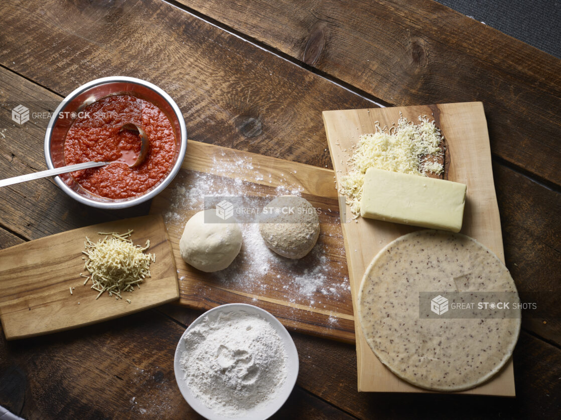 Overhead view of pizza ingredients - gluten-free crust, white and whole wheat pizza dough balls, shredded cheese, pizza sauce in a bowl with ladle, bowl of flour