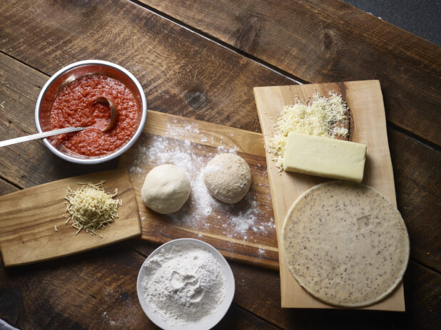Overhead view of pizza ingredients - gluten-free crust, white and whole wheat pizza dough balls, shredded cheese, pizza sauce in a bowl with ladle, bowl of flour