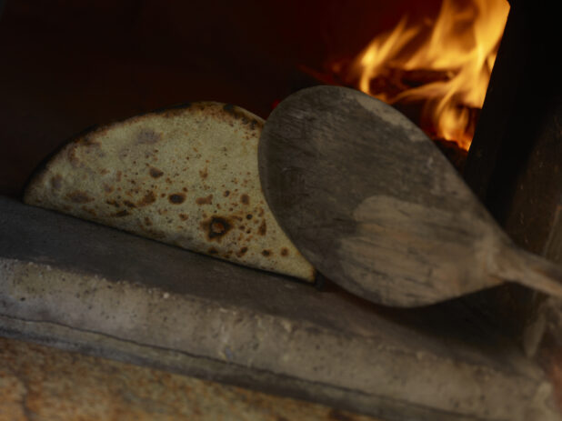 Flatbread Being Fire-Baked in a Wood-Burning Oven and Flipped with a Wooden Paddle