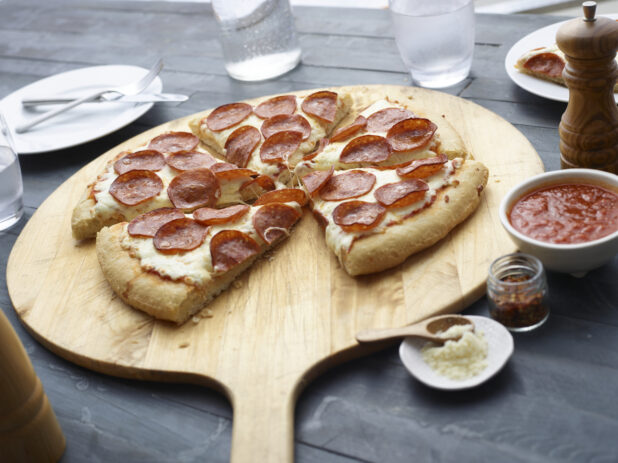 Sliced pepperoni pizza on a wood pizza peel, condiments, water glass and slice on plate alongside, all on a black wood tabletop