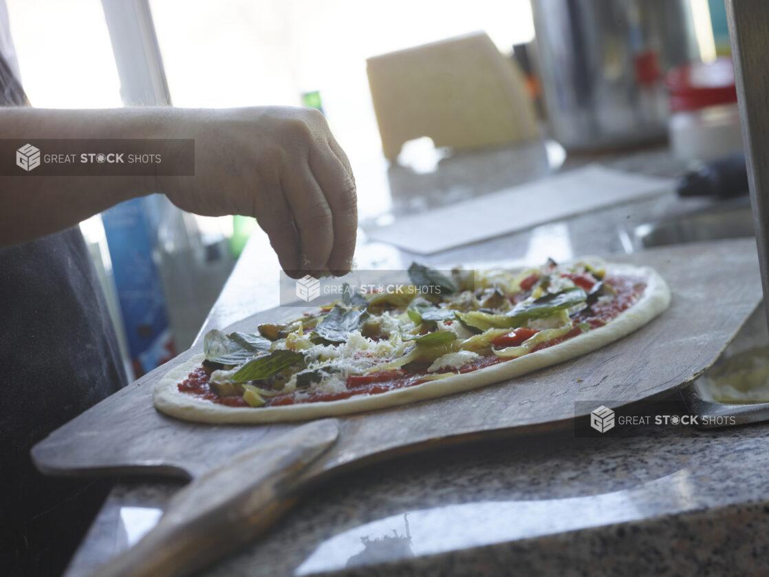 Pizza chef sprinkling ingredients on a raw pizza with grilled vegetables on a peel in a restaurant