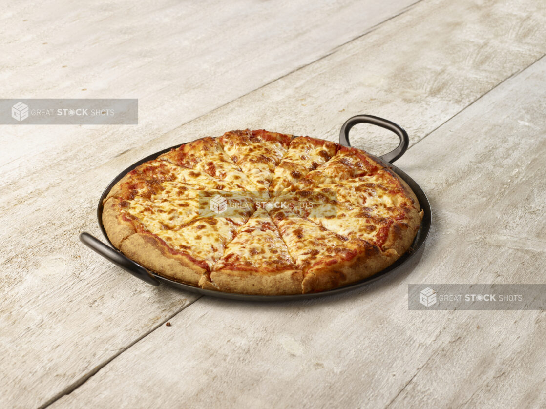 Cheese pizza in a black pan with handles on each side on a whitewashed wood background