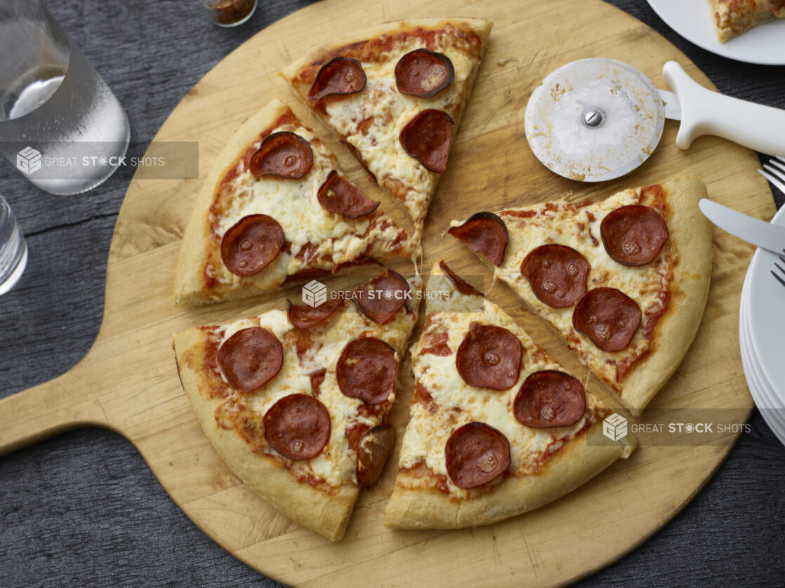 Sliced pepperoni pizza, one slice removed, on a wood pizza peel, black background, overhead view