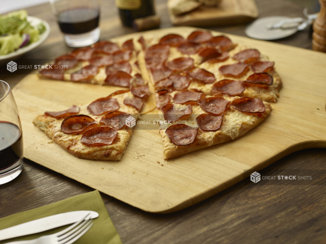 Sliced pepperoni pizza on a wood pizza peel, tumblers of red wine and place settings surrounding, low angle