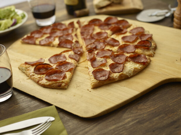 Sliced pepperoni pizza on a wood pizza peel, tumblers of red wine and place settings surrounding, low angle