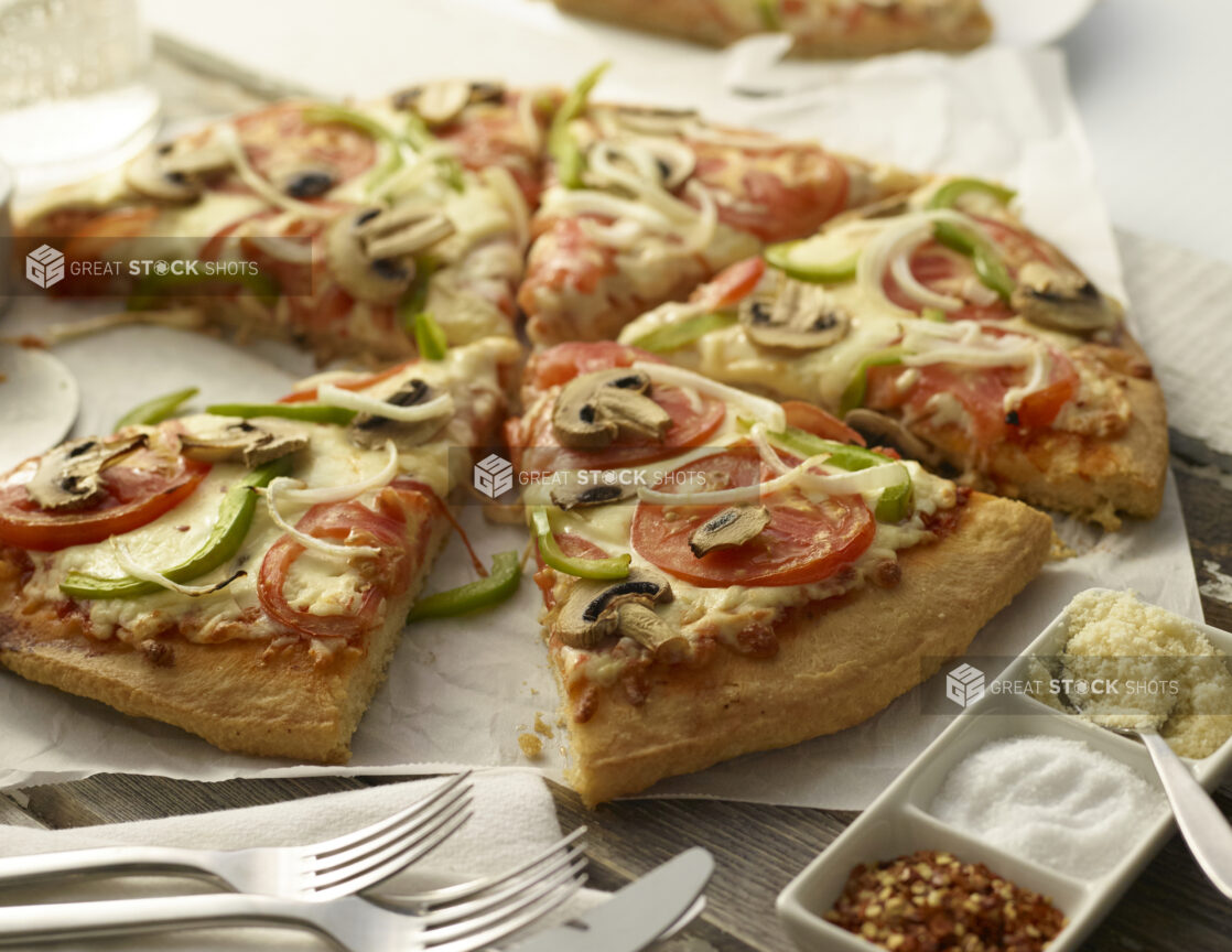 Classic deluxe vegetarian pizza, sliced, on white parchment with condiments and utensils on a wood tabletop