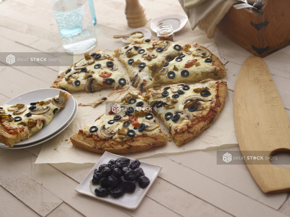 Sliced 3 topping pizza, sliced removed, on white parchment paper, dried olives and other items surrounding, wood background