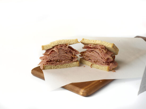 Corned beef sandwich on light rye, cut in two on white parchment on a wood cutting board