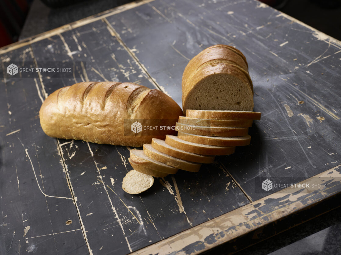 Two loaves of light rye bread, sliced and unsliced, on a black weathered wood table