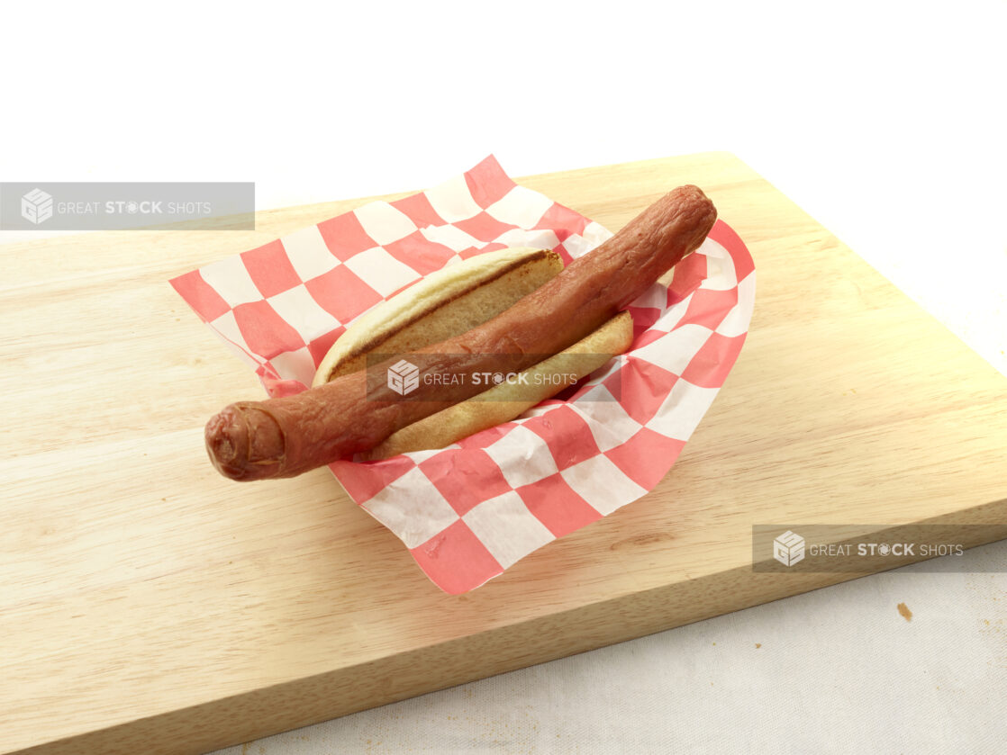 Plain foot-long hot dog on a bun on red and white checkered basket liner on a wood cutting board, light background