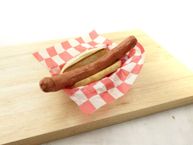 Plain foot-long hot dog on a bun on red and white checkered basket liner on a wood cutting board, light background