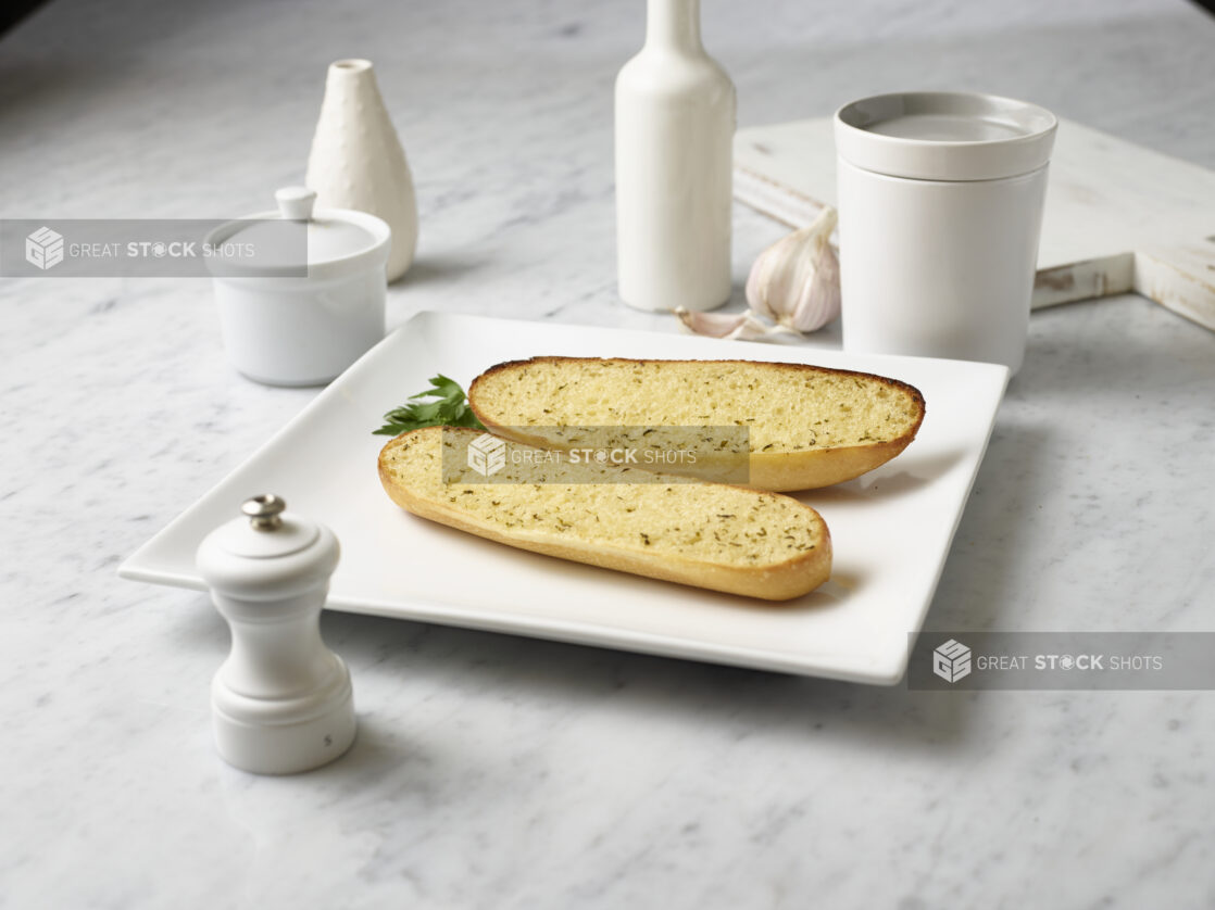 Toasted garlic bread, sliced lengthwise on a square white plate, white jars and pepper mill surrounding, white marble background