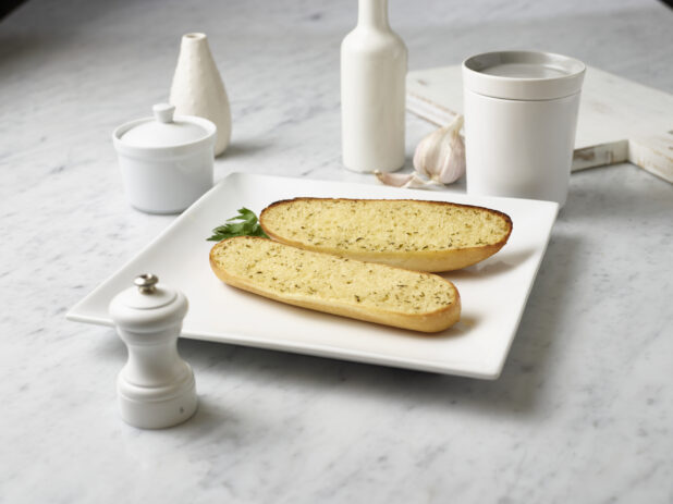 Toasted garlic bread, sliced lengthwise on a square white plate, white jars and pepper mill surrounding, white marble background