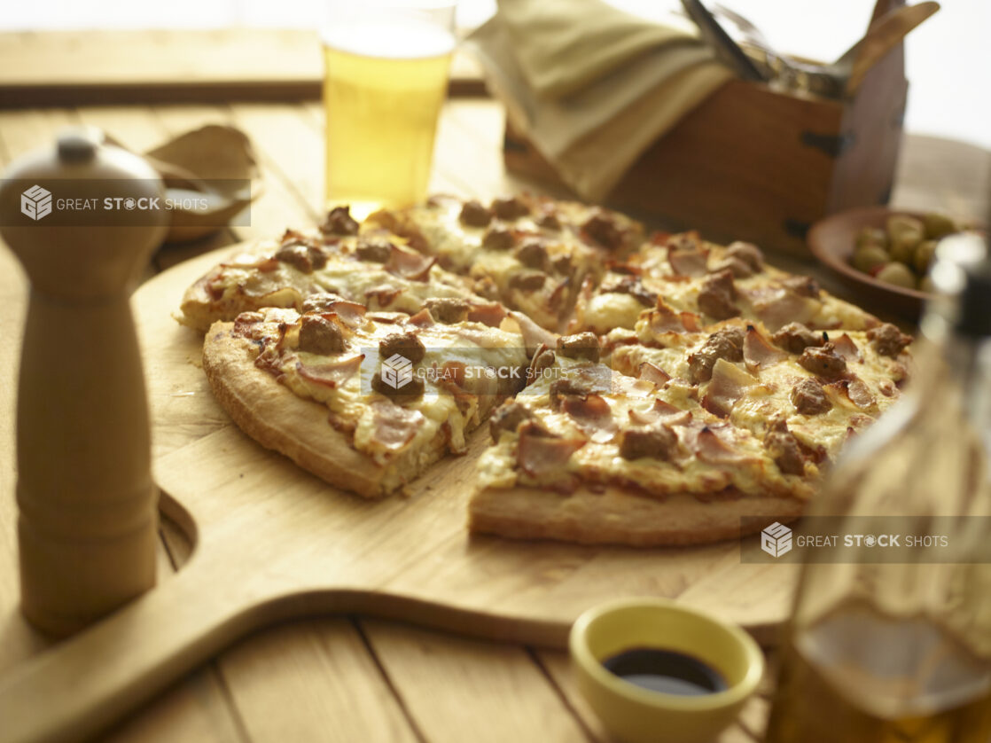 Sliced meat lovers pizza on a wood pizza peel, condiments and tableware surrounding, wood background