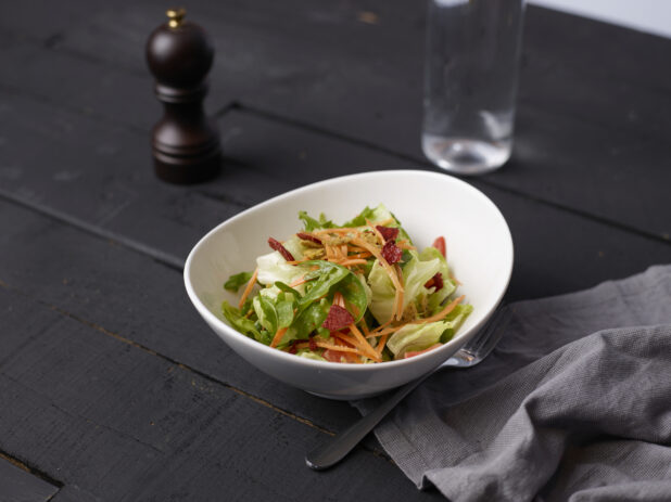 Side mesclun salad with julienned carrots and XX bits in an elegant white bowl with a fork and grey cloth napkin, dark background