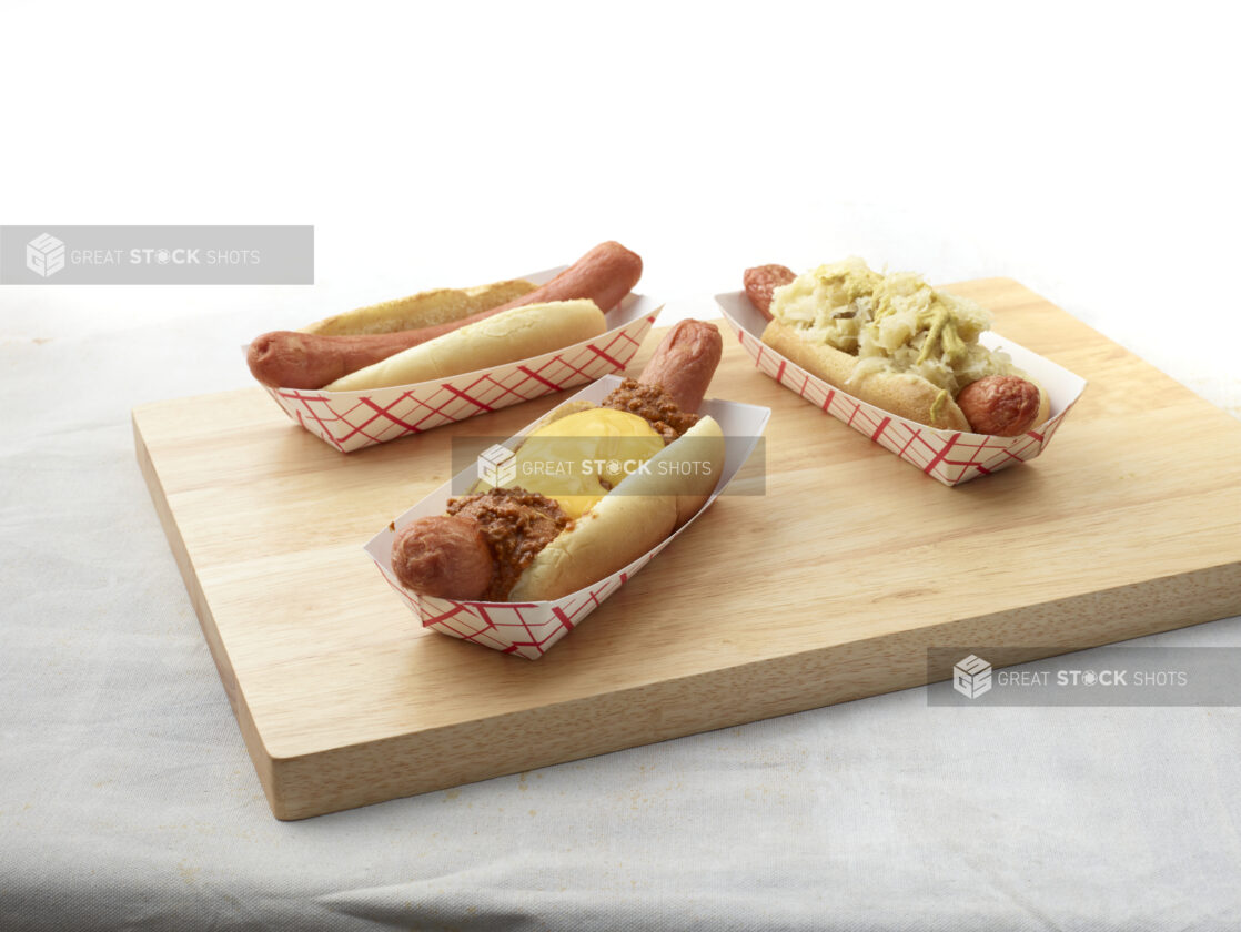 Three hot dogs with various toppings in red and white takeout containers on a wooden board, white background