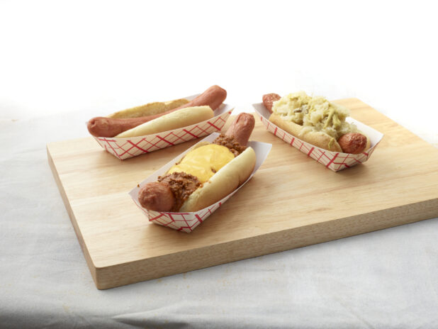 Three hot dogs with various toppings in red and white takeout containers on a wooden board, white background