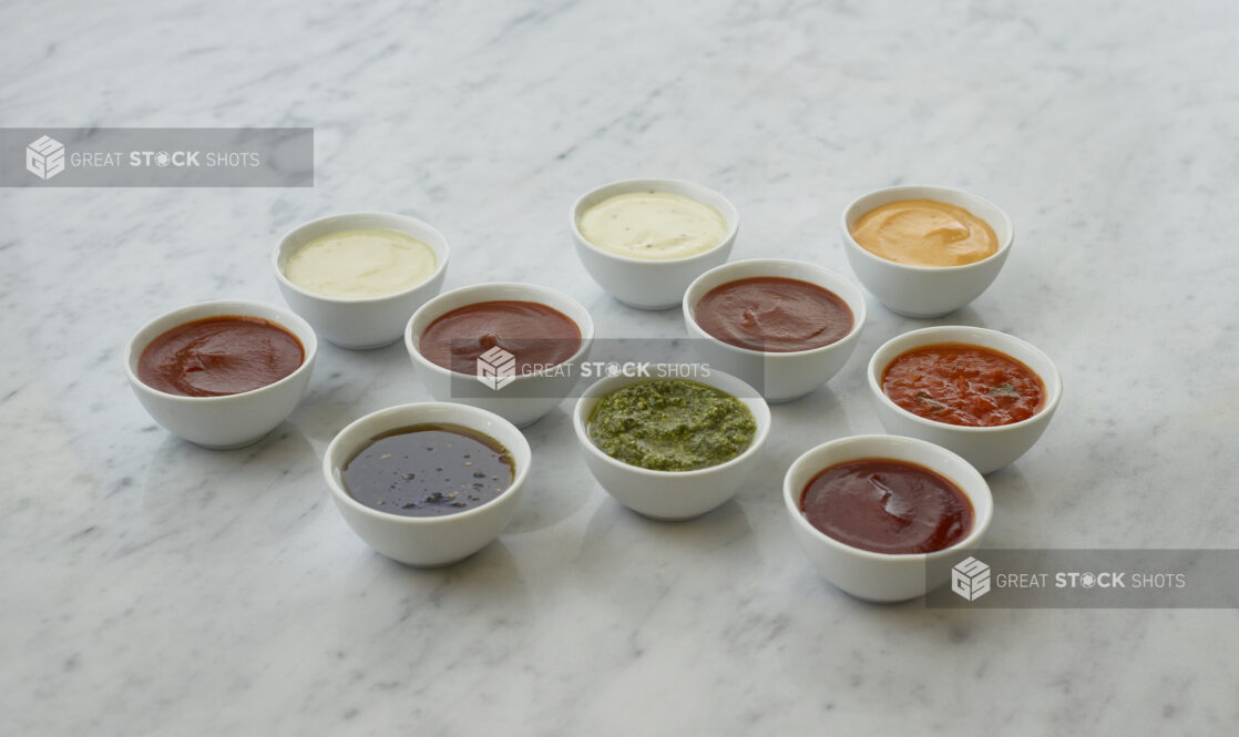 Ten round white ramekins of colourful dipping sauces and dressings, and condiments, white marble background