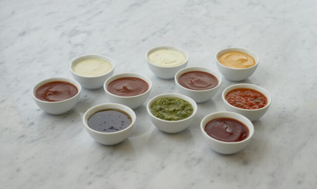 Ten round white ramekins of colourful dipping sauces and dressings, and condiments, white marble background