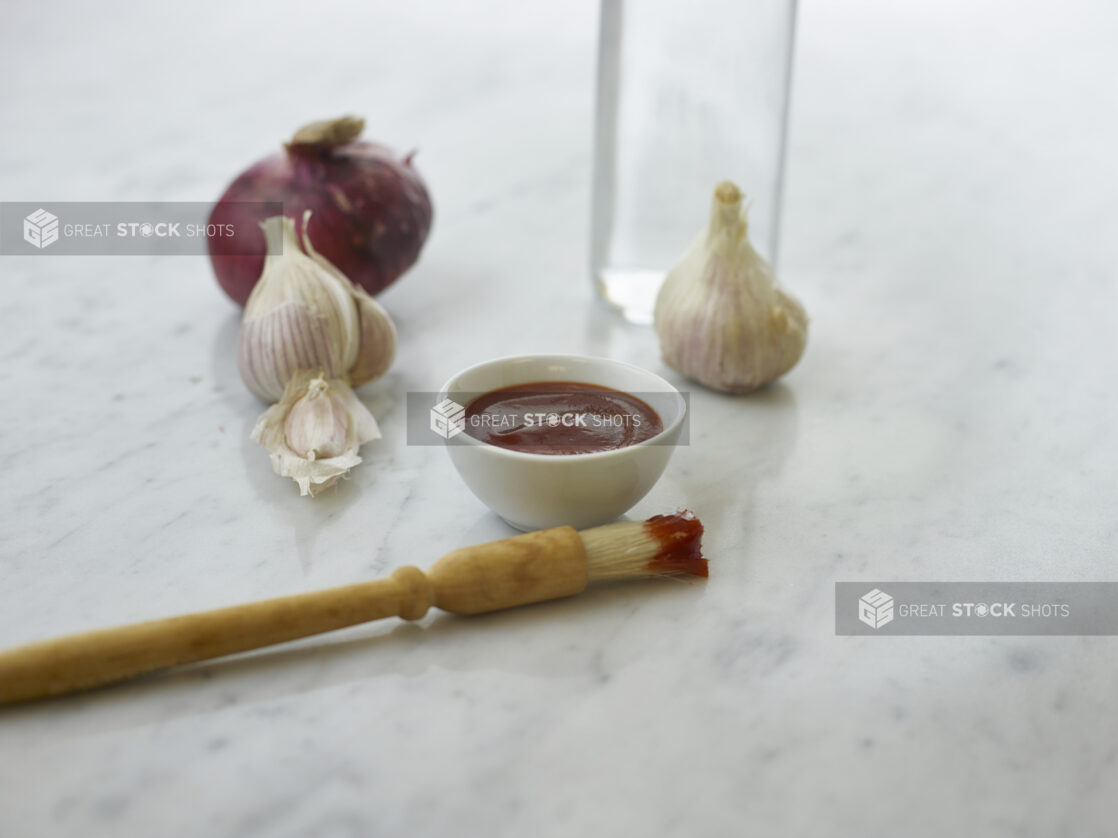 Round white ramekin of red sauce with a sauce-dipped basting brush beside it, whole garlic and onions in background, white marble background