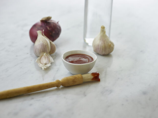 Round white ramekin of red sauce with a sauce-dipped basting brush beside it, whole garlic and onions in background, white marble background