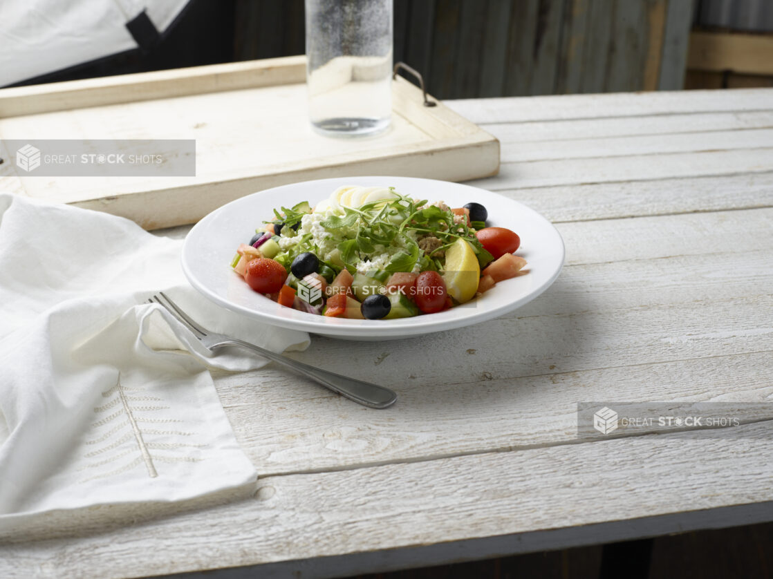 Mediterranean-style mixed green salad with arugula, feta, cherry tomatoes, black olives, boiled egg and red onion in a shallow white bowl on a whitewashed wood tabletop