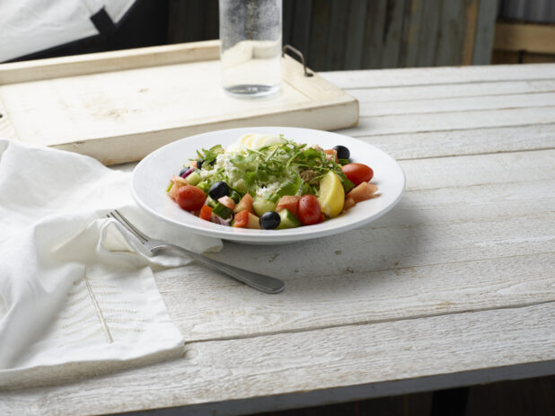 Mediterranean-style mixed green salad with arugula, feta, cherry tomatoes, black olives, boiled egg and red onion in a shallow white bowl on a whitewashed wood tabletop