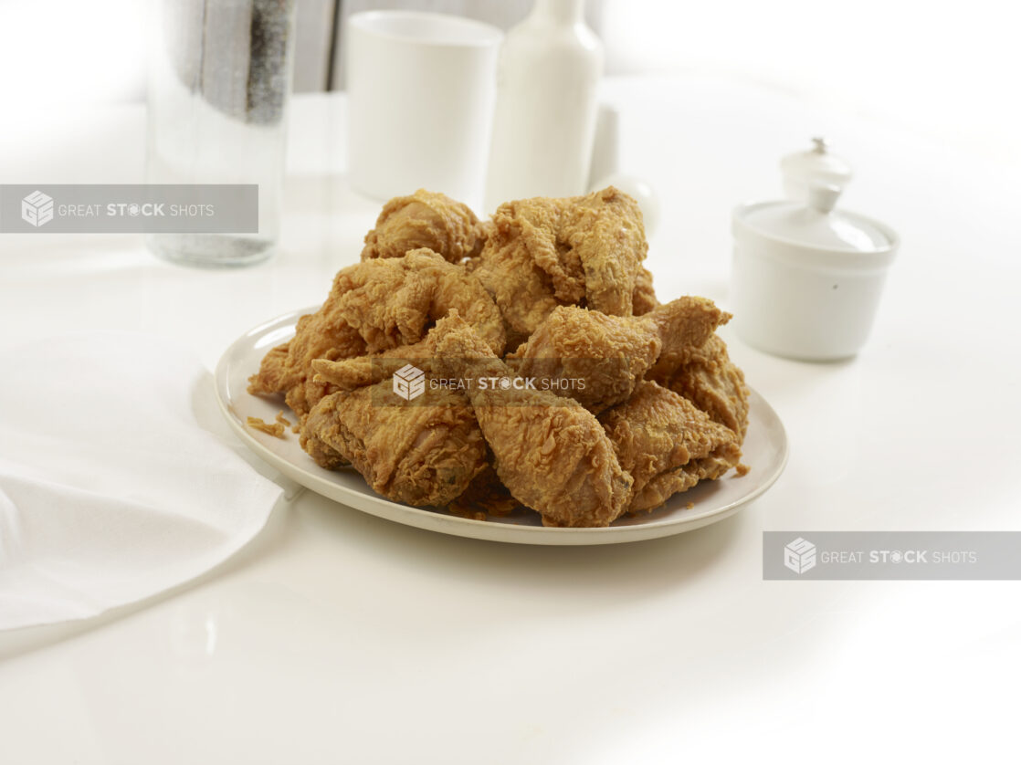 Deep fried chicken pieces piles on a round white plate, white table setting, white background