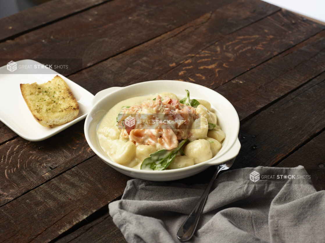 Baking dish of creamy salmon gnocchi with a side of garlic bread and cloth napkin on a dark wooden background83392