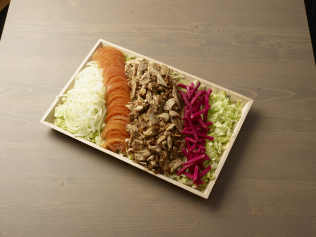 Overhead view of a wooden platter with sliced, onion, tomatoes, turnip and chicken shawarma meat on a wooden background