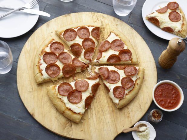 Overhead view of a sliced pepperoni pizza on wood pizza peel, one slice removed to side plate on table, dark background