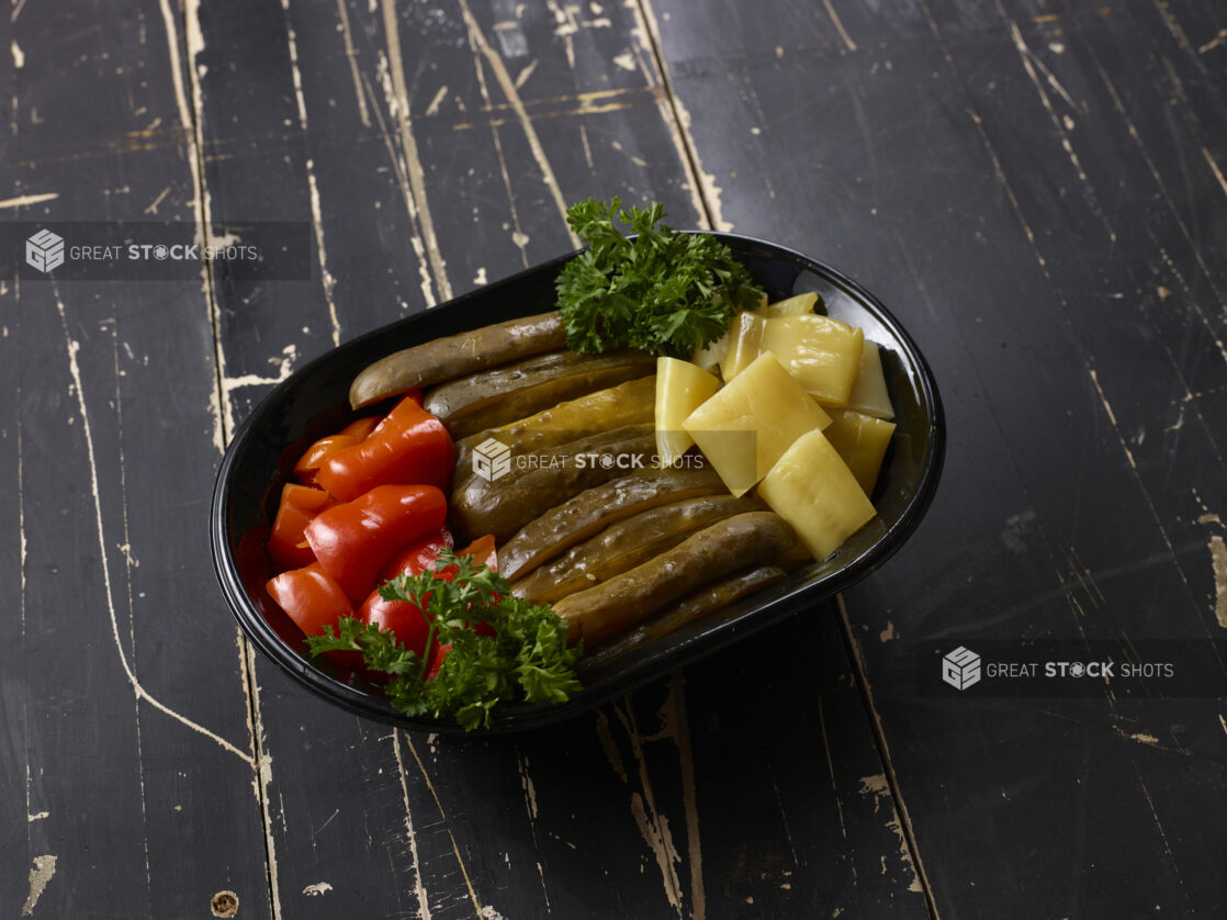 Assortment of pickled peppers and cucumbers garnished with parsley in a black oval takeout container, distressed black painted wood background