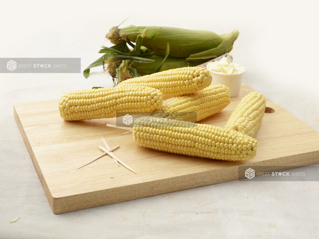 Husked cobs of fresh sweet corn piled on a wood board with toothpicks and butter, unhusked cobs in background