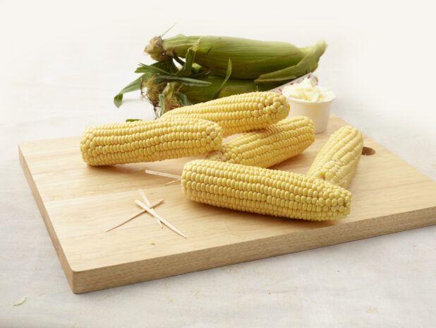 Husked cobs of fresh sweet corn piled on a wood board with toothpicks and butter, unhusked cobs in background