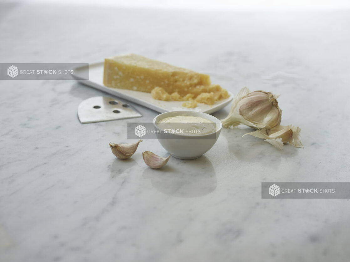 Round white ramekin of creamy dipping sauce, wedge of parmesan cheese and garlic cloves in background, white marble surface