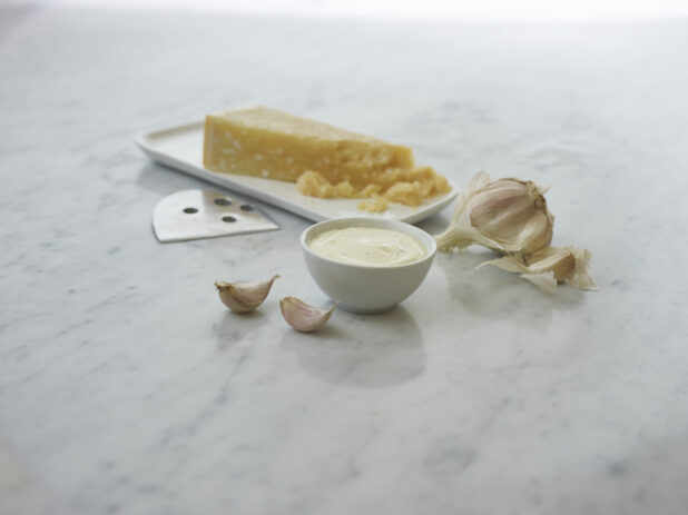 Round white ramekin of creamy dipping sauce, wedge of parmesan cheese and garlic cloves in background, white marble surface