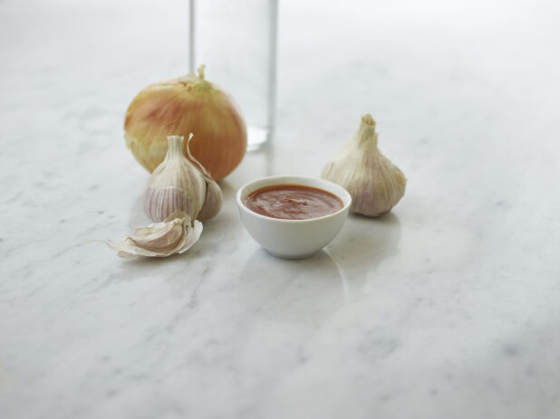 Round white ramekin of red sauce, whole garlic and onion in background, white marble background