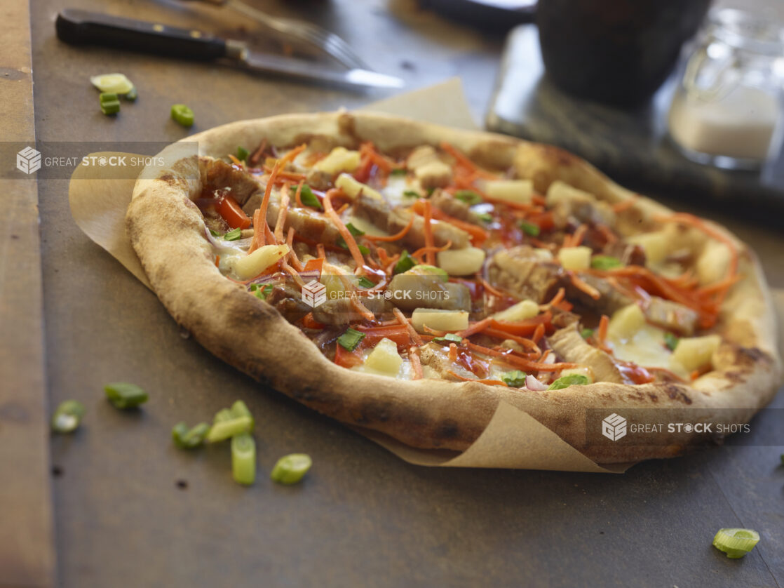 Whole pizza with pork belly, pineapple, shredded carrots, red peppers and green onions on parchment paper, close-up